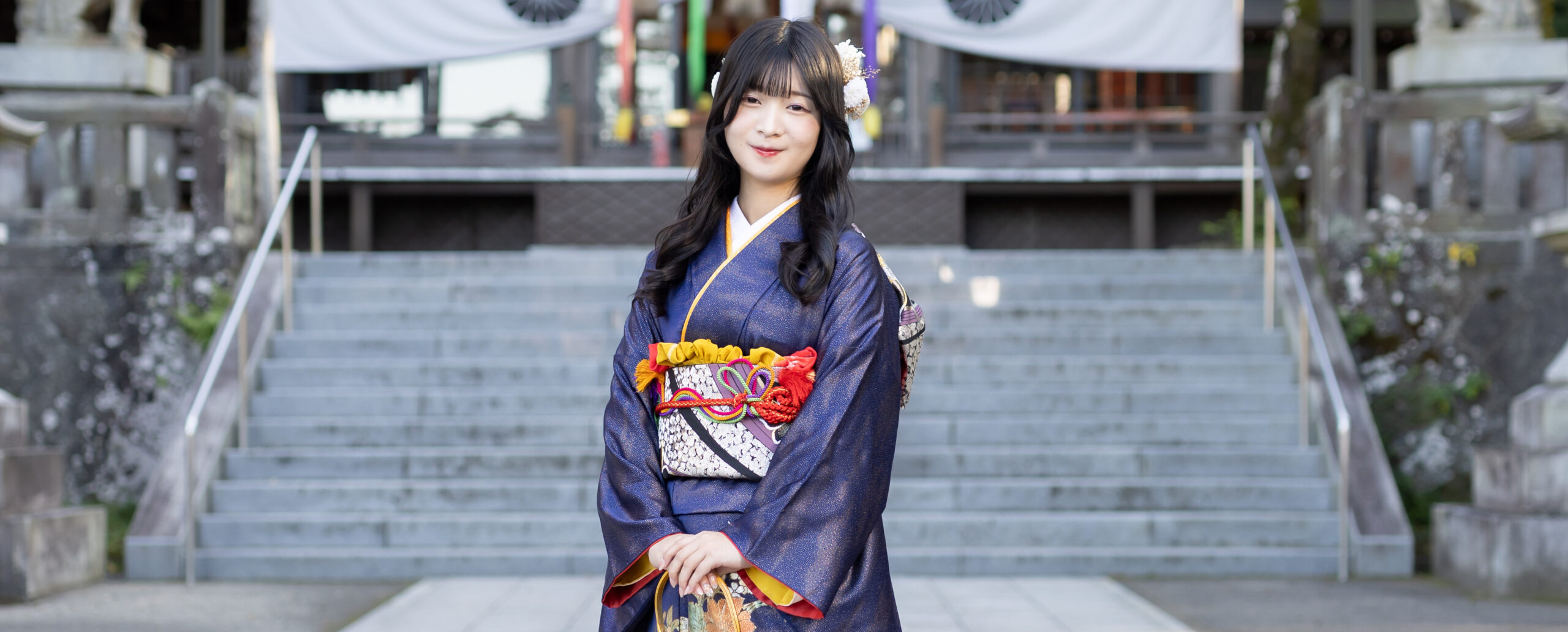 西寒田神社の石段を背景に、紺色の振袖と華やかな帯を身にまとい、やさしく微笑む女性の成人式前撮りロケーション写真
