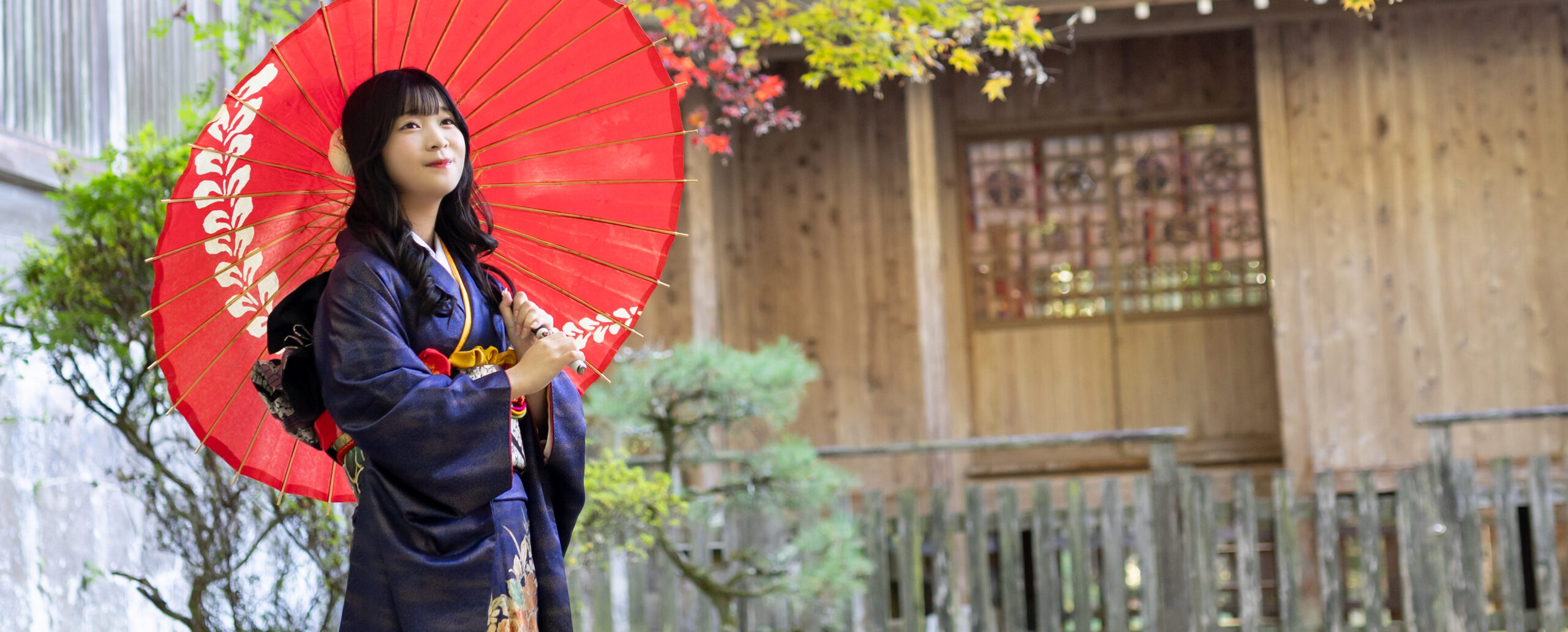 大分の西寒田神社で、紺色の振袖を着て赤い和傘を持ち、紅葉を背景に優しく微笑む女性の成人式前撮り写真